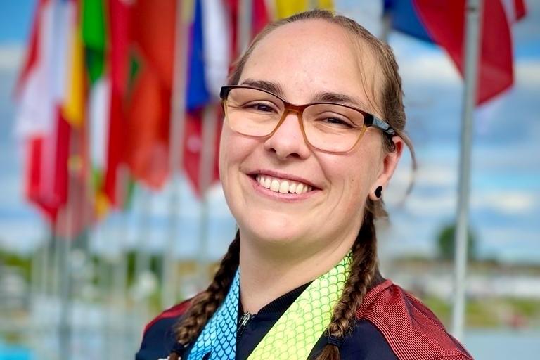 Lächelnde Athletin mit mehreren Medaillen vor einer Fahnenkulisse im Hintergrund. Blauer Himmel mit Wolken sichtbar.