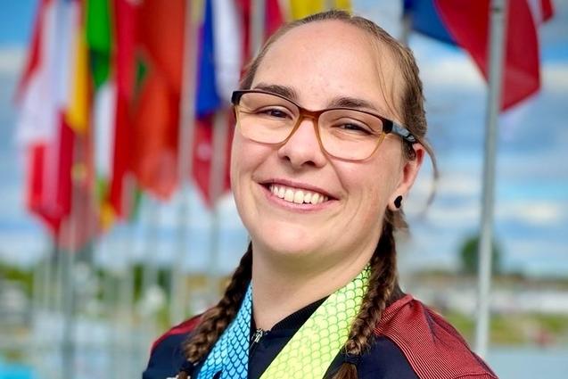 Lächelnde Athletin mit mehreren Medaillen vor einer Fahnenkulisse im Hintergrund. Blauer Himmel mit Wolken sichtbar.