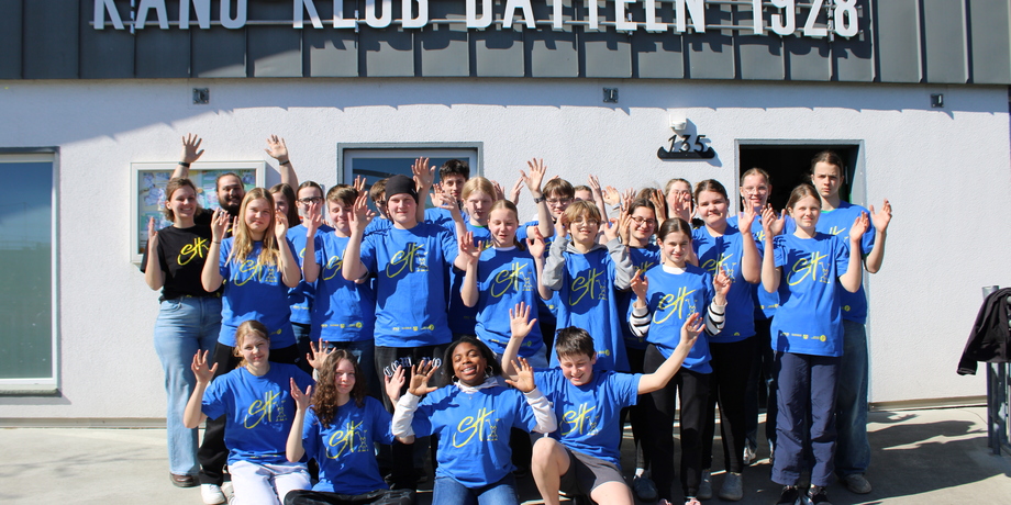 Gruppe von Jugendlichen und Erwachsenen mit erhobenen Händen vor dem Kanu-Klub Datteln, alle tragen blaue T-Shirts.