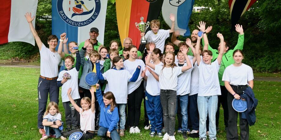 Gruppe von Jugendlichen und Erwachsenen in T-Shirts feiert mit einem Pokal vor Fahnen des Kanu-Sport-Vereins Bad Kreuznach.