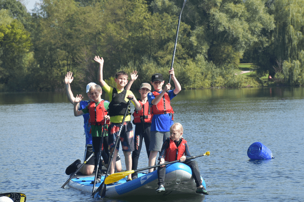 Sechs Kinder in Schwimmwesten stehen auf einem Stand-Up-Paddle-Board im Wasser und winken fröhlich.