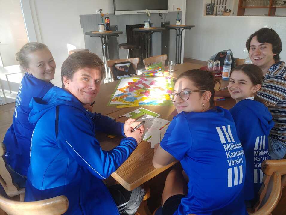 Sechs Personen sitzen um einen Tisch, beschäftigt mit bunten Karten und Spielen, alle tragen blaue Vereinsbekleidung.