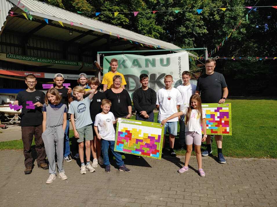 Gruppe von Jugendlichen und Erwachsenen hält bunte Pläne vor einem Kanusportgebäude im Freien.