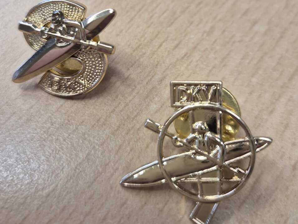 Zwei goldene Abzeichen auf einem hellen Tisch, eines mit einem Boot und das andere mit einem Paddel.
