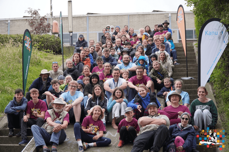 Gruppenselfie von etwa 80 Personen auf einer Treppe, umgeben von grüner Natur und Werbebannern für ein Kanu-Team.