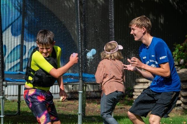 Zwei Kinder spielen auf einem Trampolin, einer spritzt Wasser, während ein anderes Kind zuschaut. Sonniger Tag im Freien.