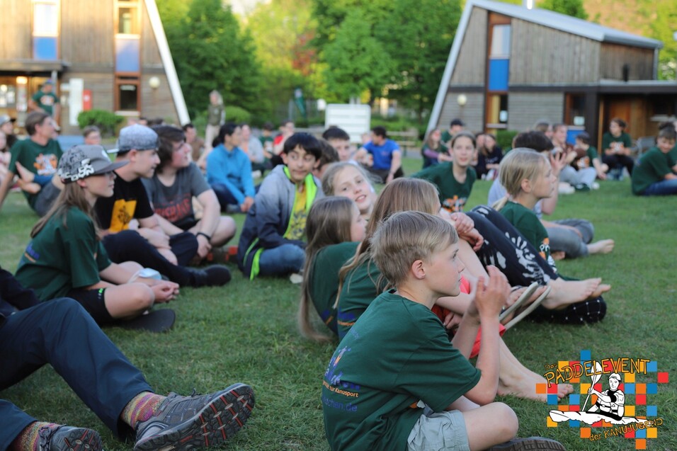 Gruppenszene mit Kindern und Jugendlichen, die auf einer Wiese vor modernen Hütten sitzen und aufmerksam etwas verfolgen.