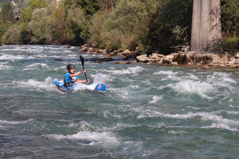 Junger Kajakfahrer paddelt durch einen wilden Fluss mit steinigen Ufern und bewaldeten Hängen im Hintergrund.