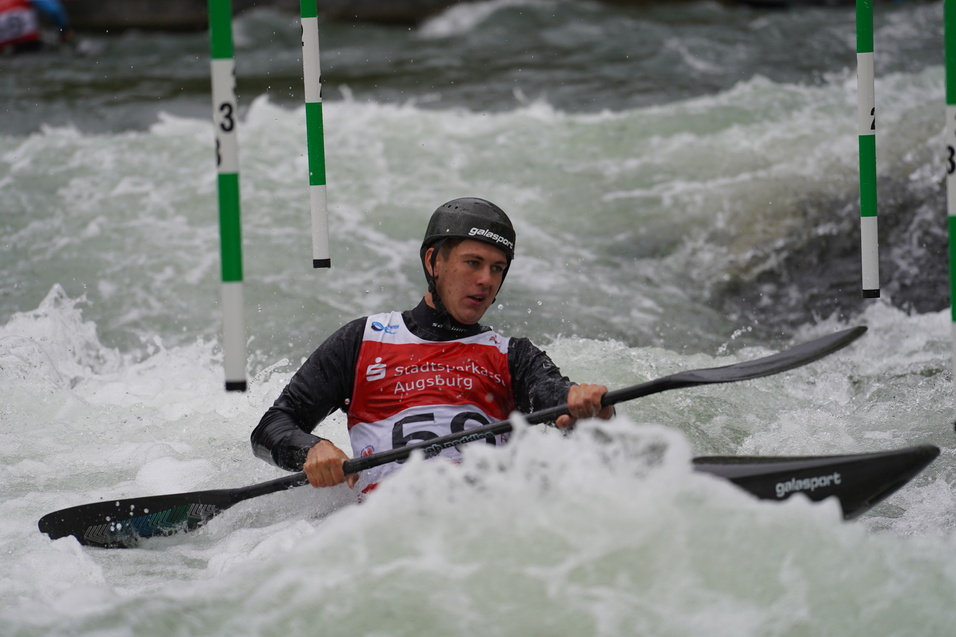 Sportler im Kanu paddelt durch sprudelndes Wasser und navigiert zwischen den grünen Torstangen im Wildwasser.