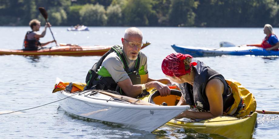 Zwei Kajakfahrer im Wasser, einer hilft der anderen, während im Hintergrund weitere Kajaks fahren.