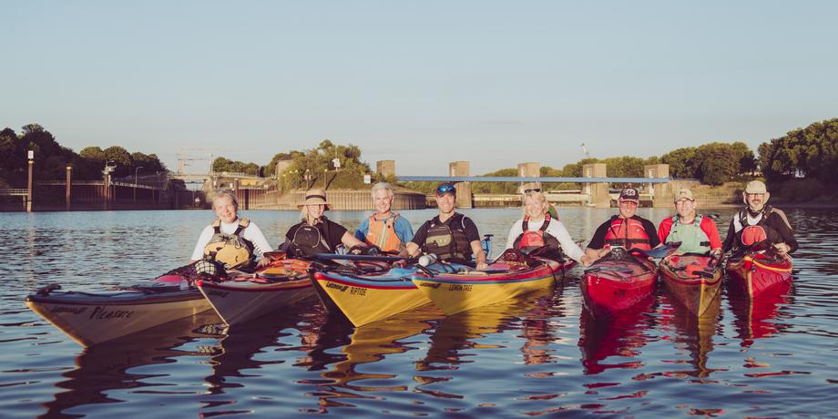 Sechs Kajakfahrer posieren in unterschiedlichen Kajaks auf einem ruhigen Gewässer vor einer Schleuse.