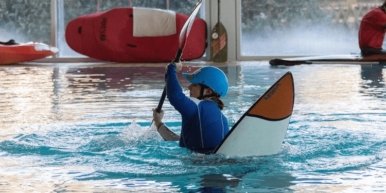 Person im Wasser beim Kajakfahren, trägt einen Helm und paddelt mit einem Paddel in einem Innenbecken.