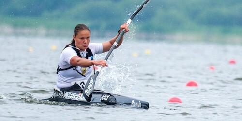 Sportlerin paddelt mit einem Kanu auf einem Gewässer, umgeben von roten Bojen und einer grünen Landschaft im Hintergrund.
