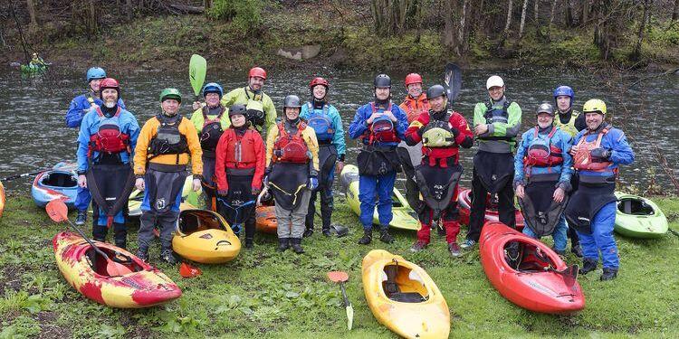 Gruppe von Paddlern in Neoprenanzügen neben bunten Kajaks an einem Flussufer.