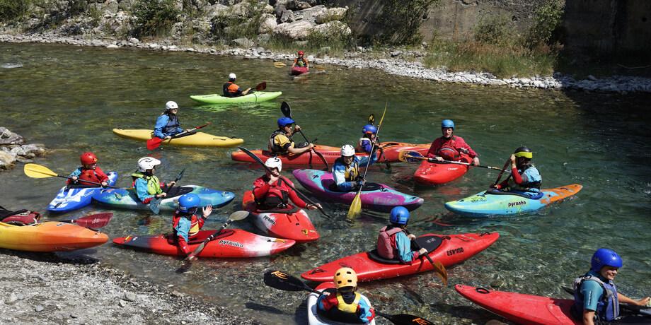 Gruppierung von Kajakfahrern in bunten Booten auf einem klaren Fluss, umgeben von Ufersteinen und Vegetation.