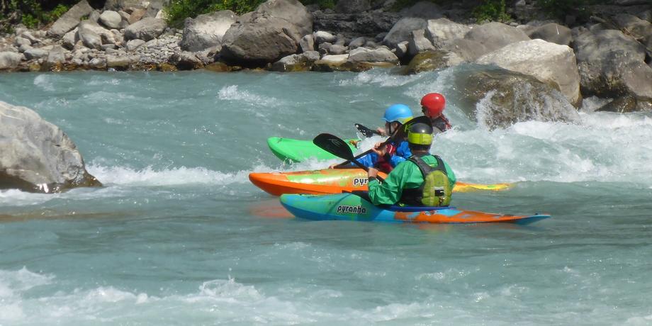 Drei Kajakfahrer paddeln auf einem wild fließenden Fluss zwischen großen Steinen bei sonnigem Wetter.