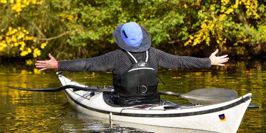 Person im Kajak mit ausgebreiteten Armen auf ruhigem Gewässer, umgeben von herbstlichen Bäumen mit gelben Blättern.