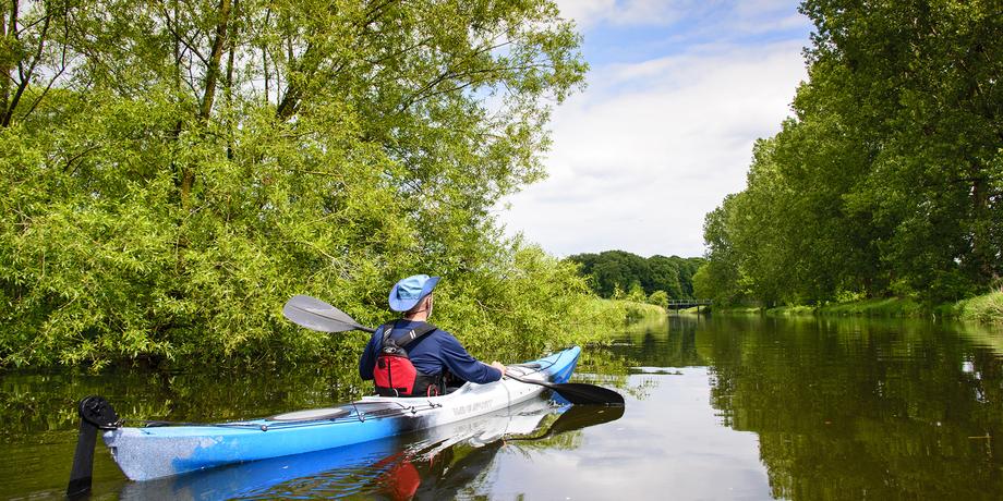 Person im Kajak paddelt auf ruhigem Wasser zwischen grünen Bäumen unter einem teilweise bewölkten Himmel.