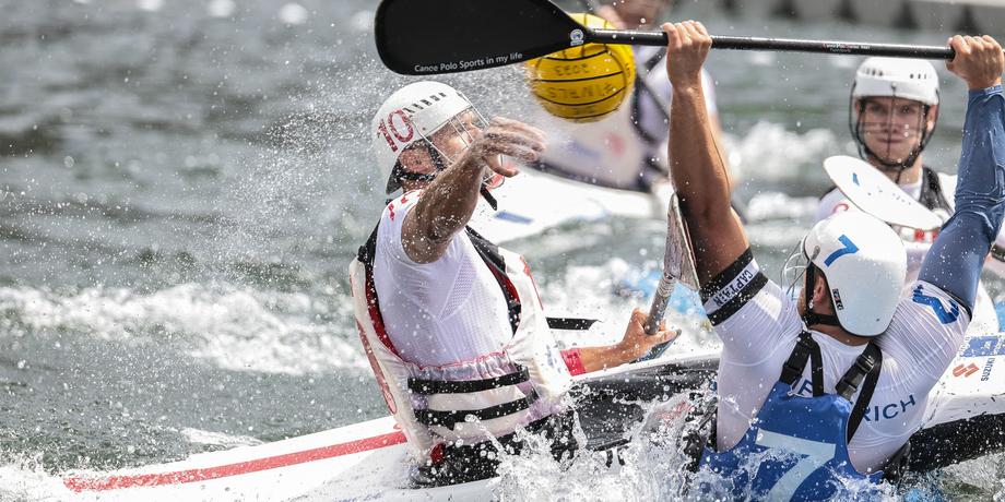 Zwei Kanupolo-Spieler in einem Wettkampf, spritzendes Wasser und ein gelber Ball sind sichtbar.