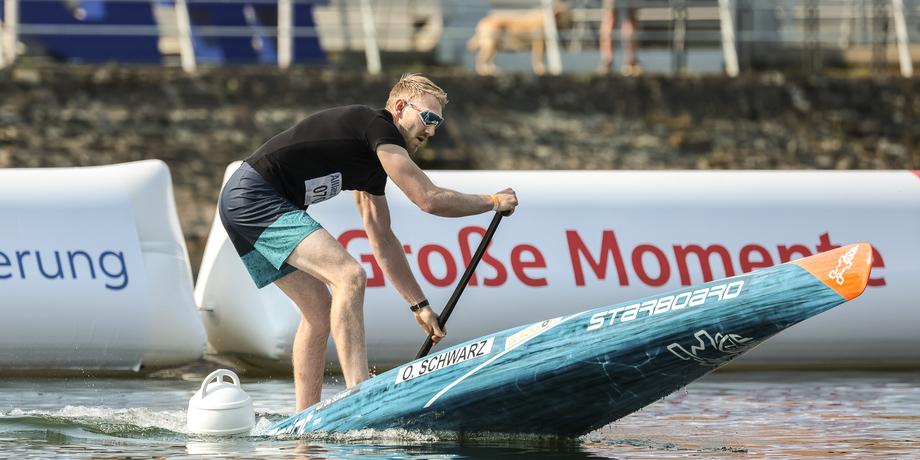 Ein Mann paddelt auf einem blauen Stand-Up-Paddleboard in einem Wettkampf, umgeben von Wasser und Bannern.