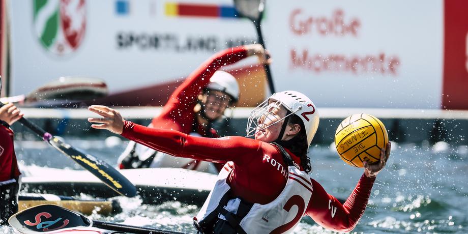 Eine Spielerin wirft einen Wasserball aus einem Kanu, wobei Wasser spritzt, im Hintergrund weitere Kanufahrer.