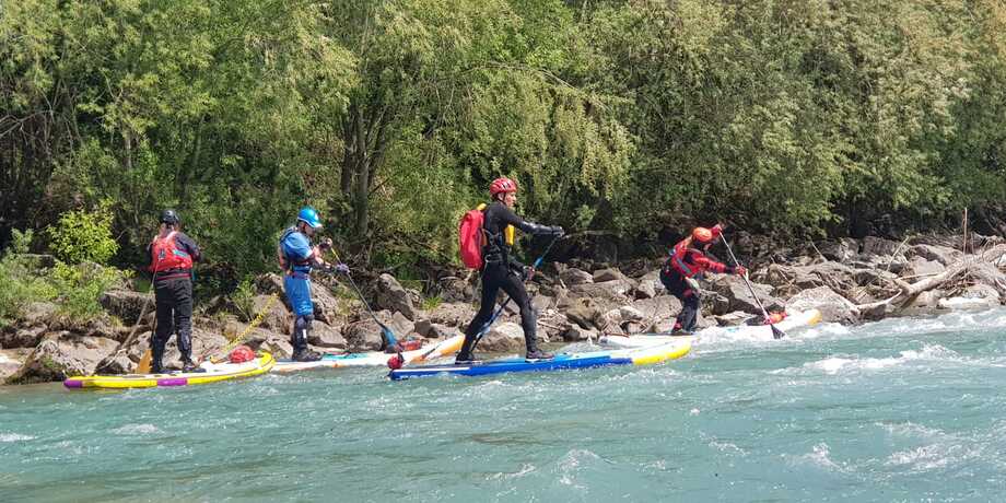 Gruppenerlebnis beim Stand-Up-Paddling auf einem Fluss, mit Sportlern in Sicherheitsausrüstung auf Board und im Wasser.