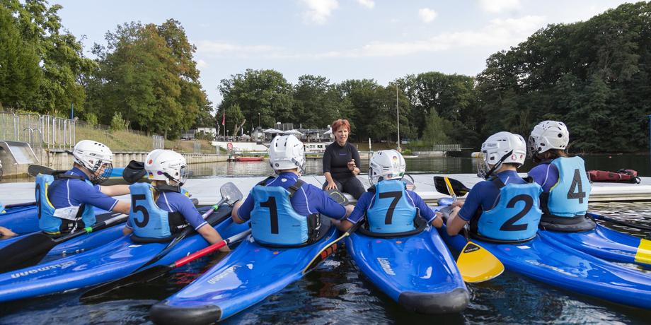 Gruppentraining im Kanu mit sechs Teilnehmern und einem Trainer am Wasser, umgeben von Bäumen und einer Freizeitanlage.