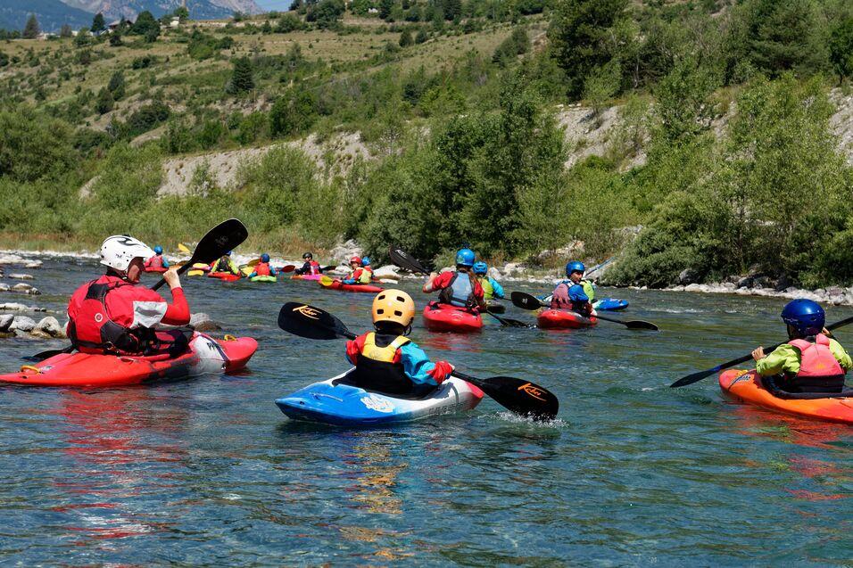 Gruppe von Kajakfahrern paddelt auf einem klaren Fluss vor einer grünen Uferlandschaft unter blauem Himmel.