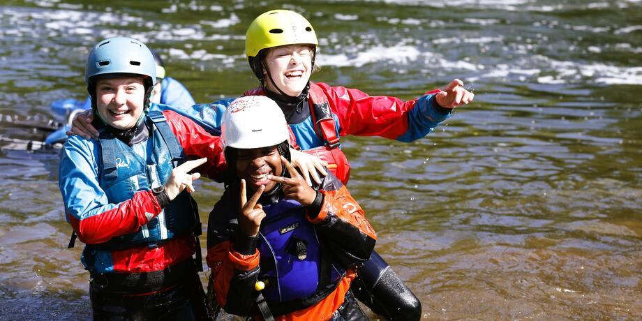 Drei Jugendliche in Neoprenanzügen halten sich im Wasser und lächeln fröhlich, während sie gemeinsam posieren.