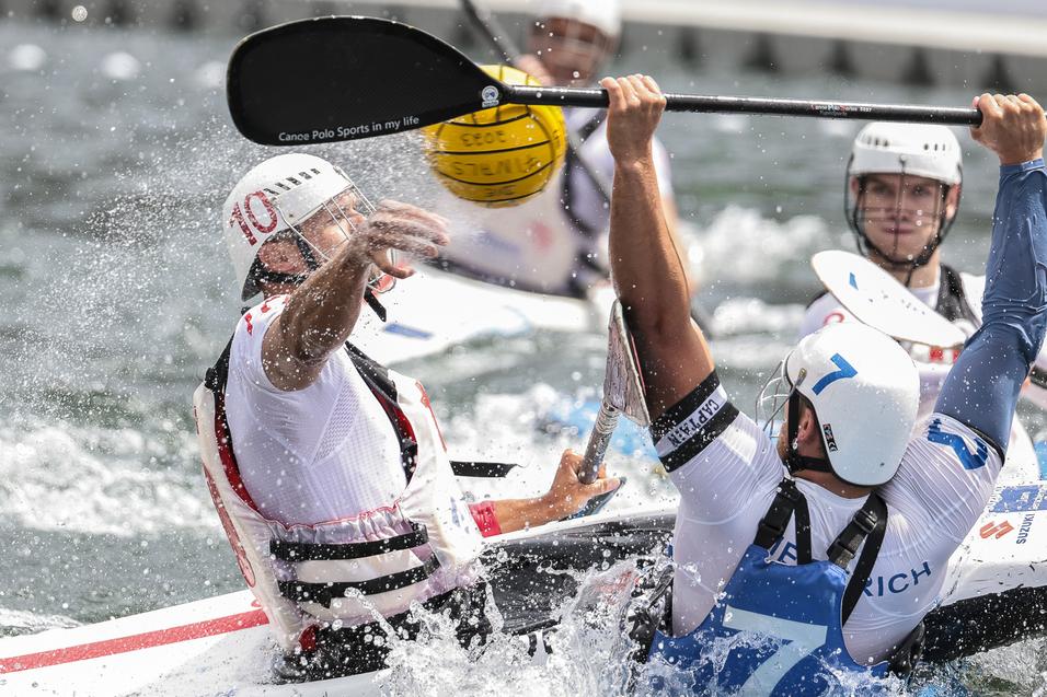 Zwei Kanupolo-Spieler in einem Wettkampf, spritzendes Wasser und ein gelber Ball sind sichtbar.