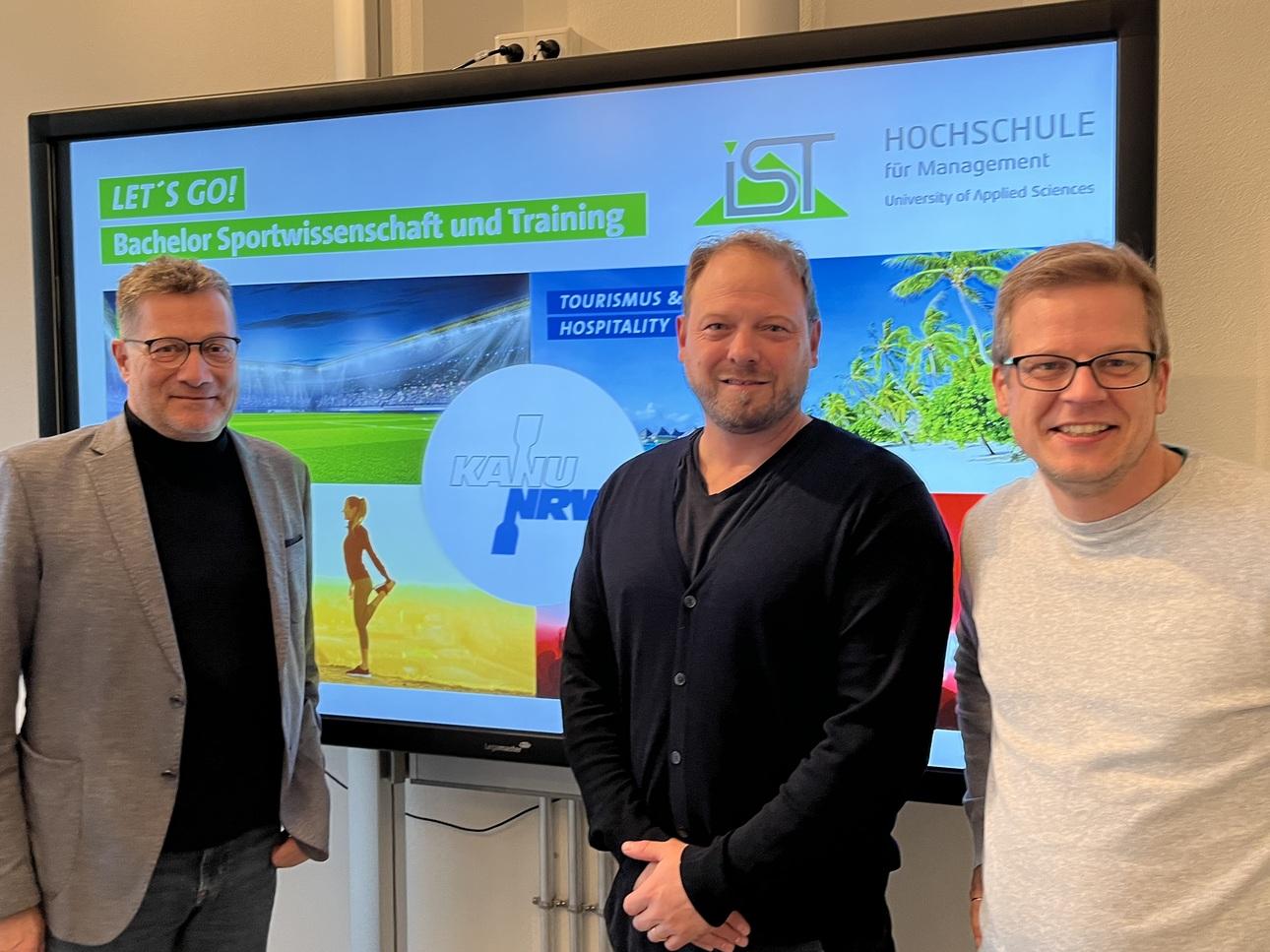 Drei Männer stehen vor einer Präsentation mit dem Titel „Bachelor Sportwissenschaft und Training“ und weiteren grafischen Elementen.