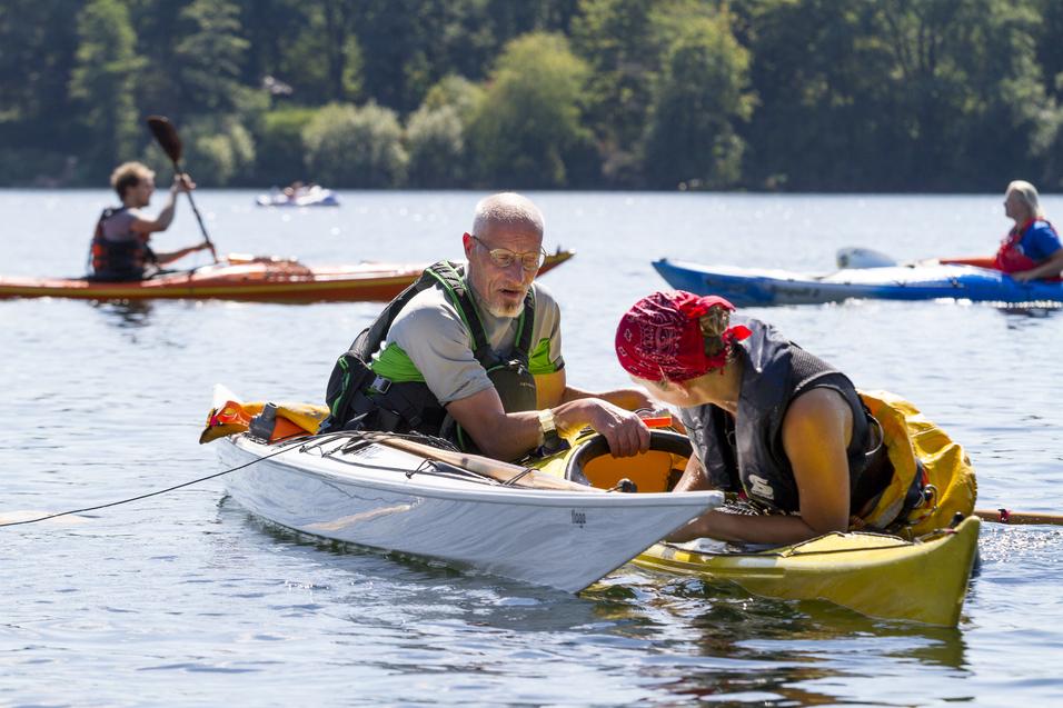 Zwei Kajakfahrer im Wasser, einer hilft der anderen, während im Hintergrund weitere Kajaks fahren.