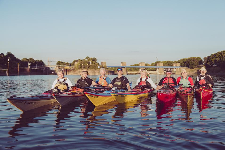 Sechs Kajakfahrer posieren in unterschiedlichen Kajaks auf einem ruhigen Gewässer vor einer Schleuse.