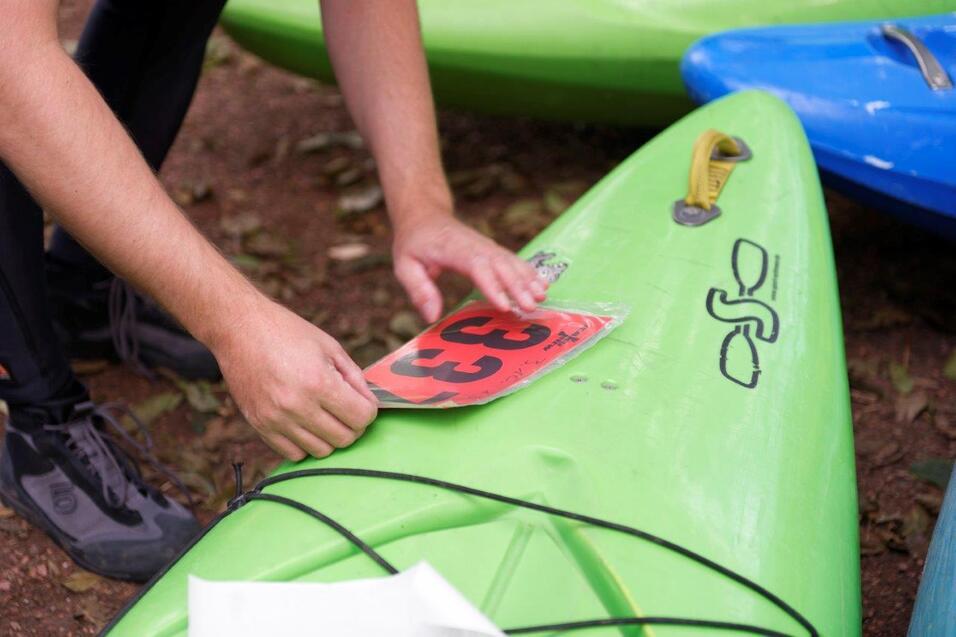 Eine Person bringt eine orangefarbene Nummer 33 auf einem grünen Kajak an, das im Freien steht.