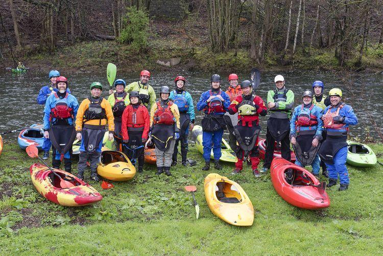 Gruppe von Paddlern in Neoprenanzügen neben bunten Kajaks an einem Flussufer.