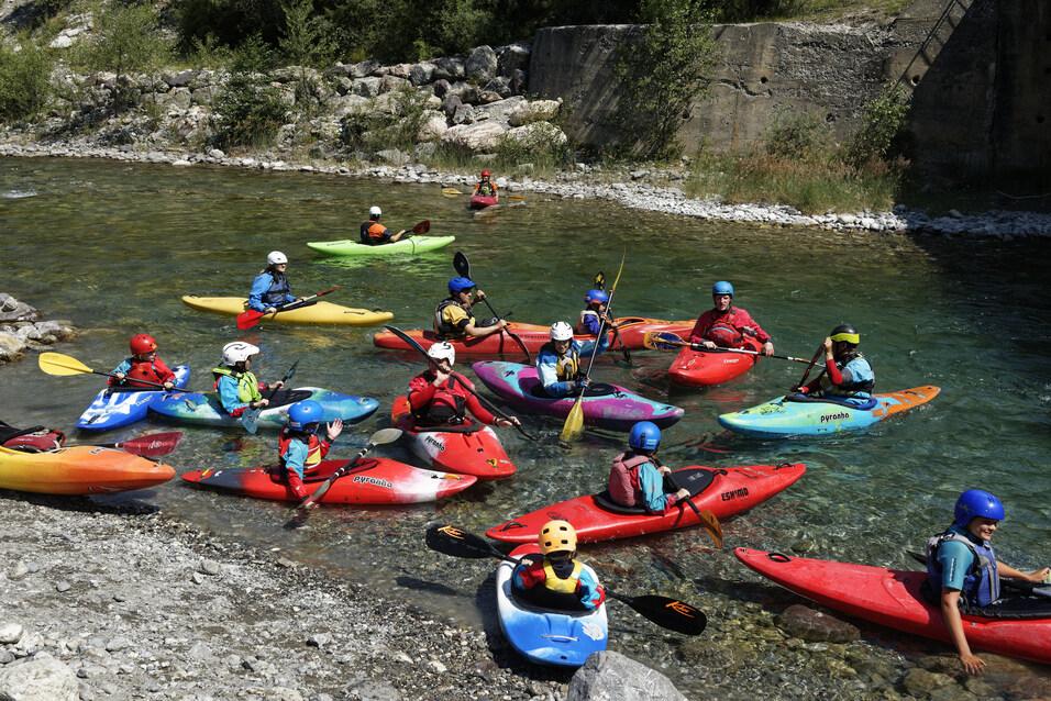 Gruppierung von Kajakfahrern in bunten Booten auf einem klaren Fluss, umgeben von Ufersteinen und Vegetation.