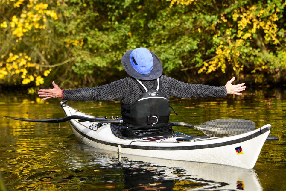 Person im Kajak mit ausgebreiteten Armen auf ruhigem Gewässer, umgeben von herbstlichen Bäumen mit gelben Blättern.