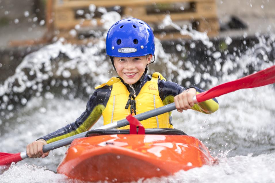 Junge im orangefarbenen Kajak paddelt mit rotem Paddel durch sprudelndes Wasser, trägt blaue Schutzkleidung.