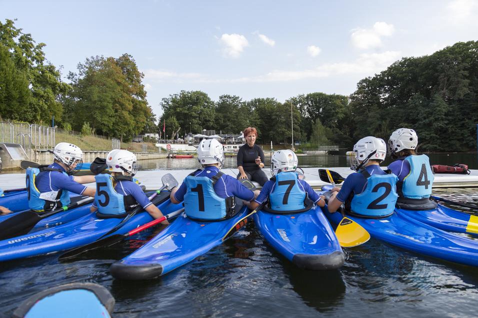 Gruppentraining im Kanu mit sechs Teilnehmern und einem Trainer am Wasser, umgeben von Bäumen und einer Freizeitanlage.