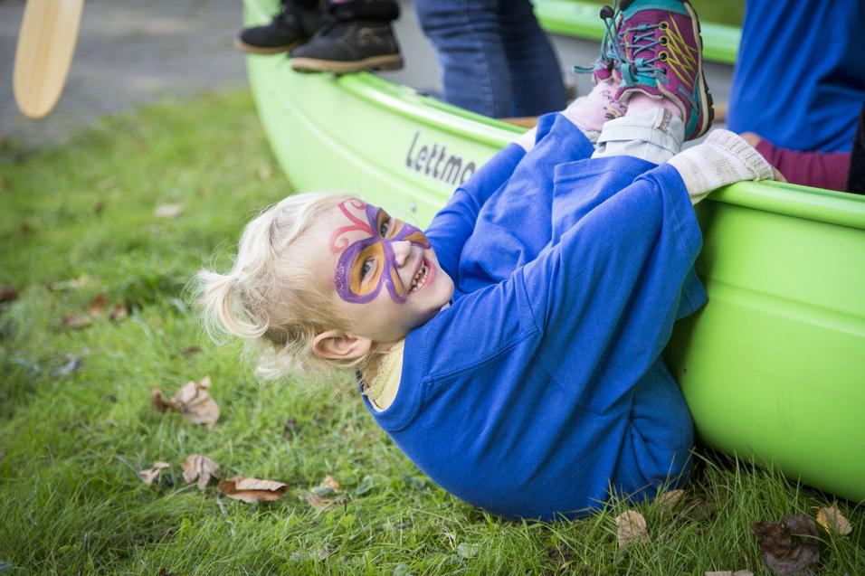 Ein Kind mit Gesichtsbemalung lächelt und sitzt am Ufer eines grünen Boots, umgeben von Gras und Laub.