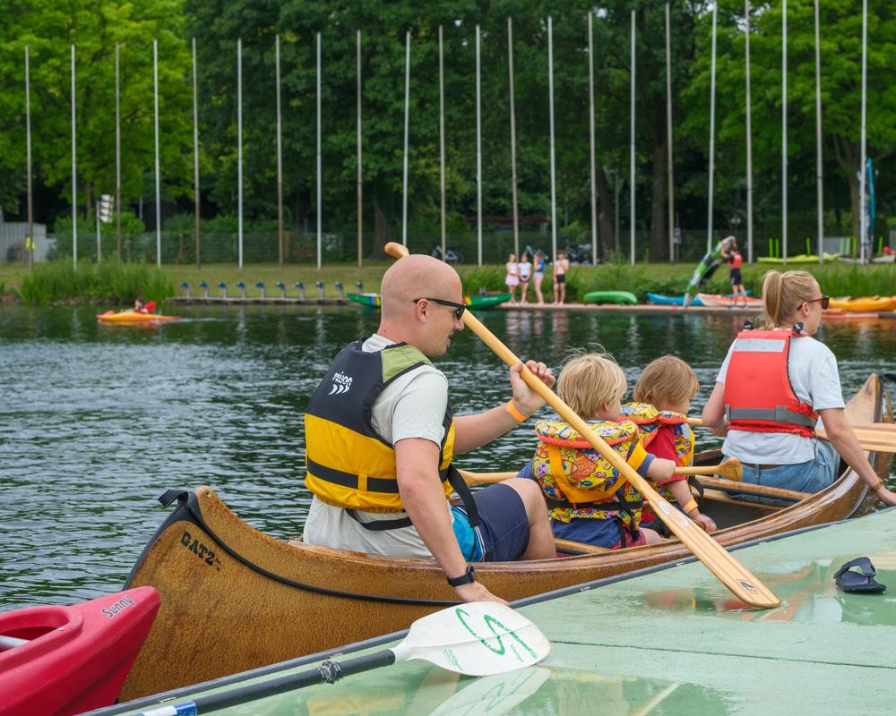 Familie im Kanu, zwei Kinder mit Schwimmwesten, paddeln auf einem See. Im Hintergrund stehen weitere Boote.