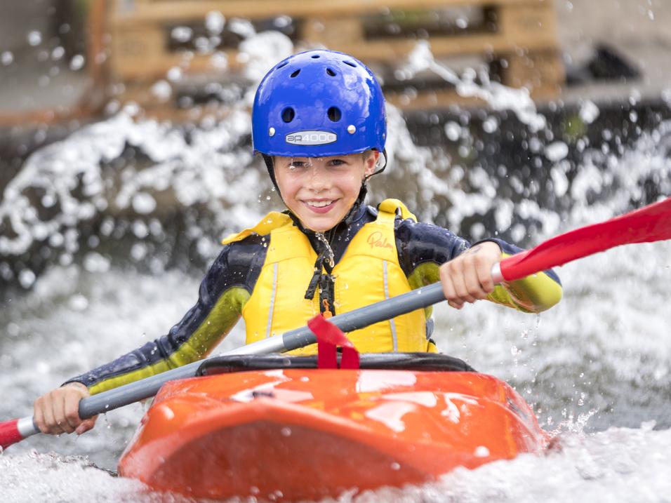 Junge im orangefarbenen Kajak paddelt mit rotem Paddel durch sprudelndes Wasser, trägt blaue Schutzkleidung.