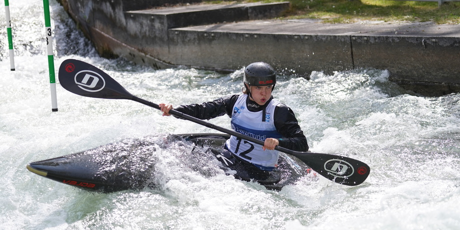 Kanute paddelt konzentriert in einem Wildwasserkanal, während er grüne Tore umschifft. Spritzwasser umgibt das Boot.