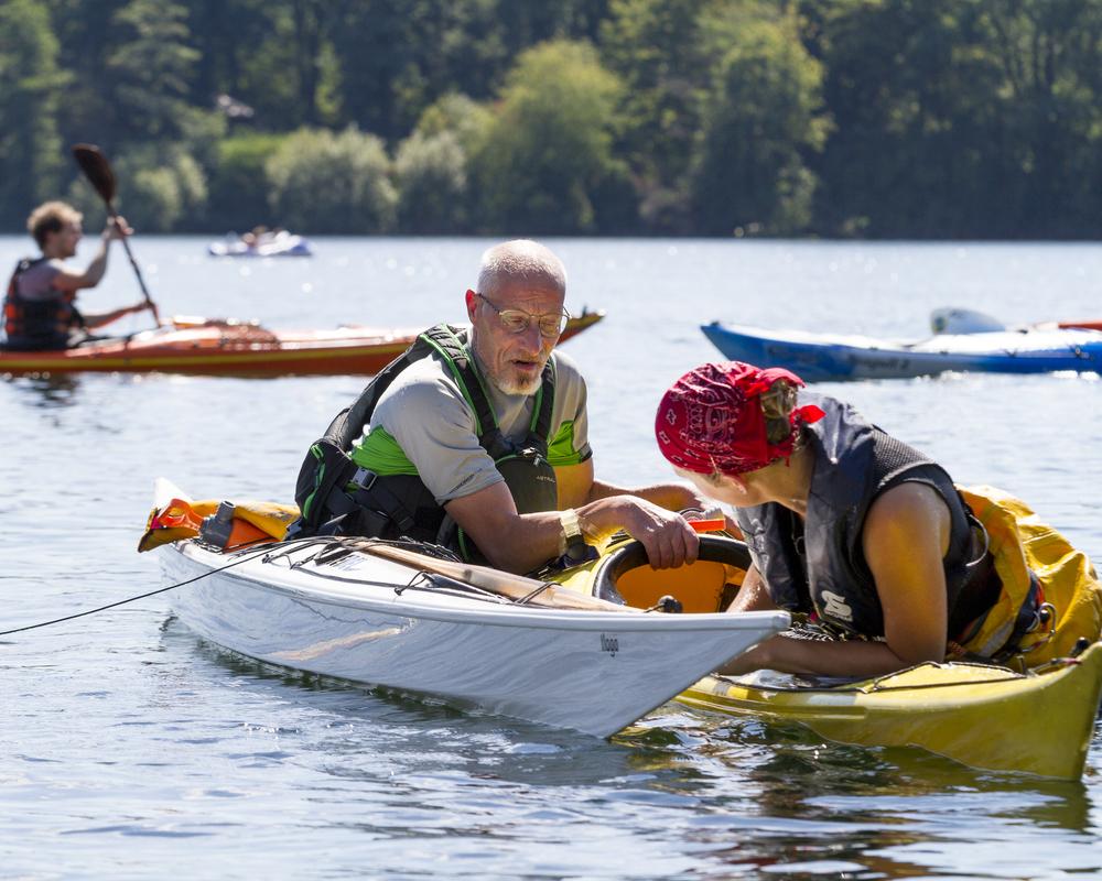 Zwei Kajakfahrer im Wasser, einer hilft der anderen, während im Hintergrund weitere Kajaks fahren.