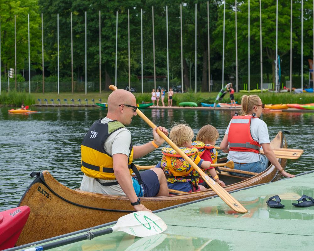 Familie im Kanu, zwei Kinder mit Schwimmwesten, paddeln auf einem See. Im Hintergrund stehen weitere Boote.