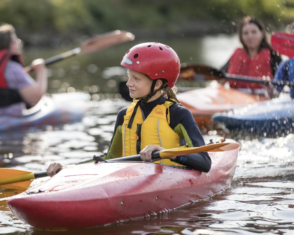 Ein Mädchen in einem roten Kajak trägt einen roten Helm und eine gelbe Schwimmweste, umgeben von weiteren Kajakfahrern.