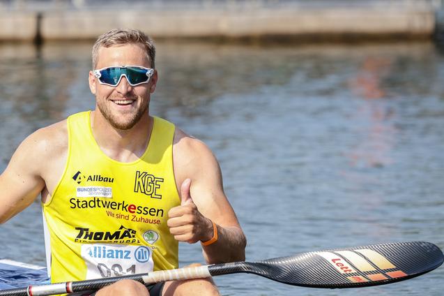 Lächelnder Kanufahrer in gelbem Trikot sitzt im Boot und zeigt Daumen hoch vor einem Gewässer.