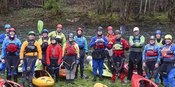 Gruppe von Paddlern in Neoprenanzügen neben bunten Kajaks an einem Flussufer.