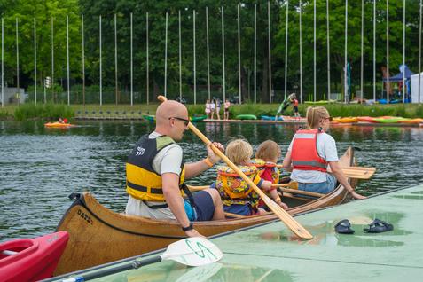 Familie im Kanu, zwei Kinder mit Schwimmwesten, paddeln auf einem See. Im Hintergrund stehen weitere Boote.