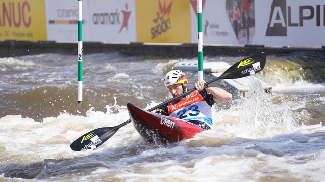 Kanufahrer im Wettkampf, paddelt durch schäumendes Wasser mit Torstangen im Hintergrund.